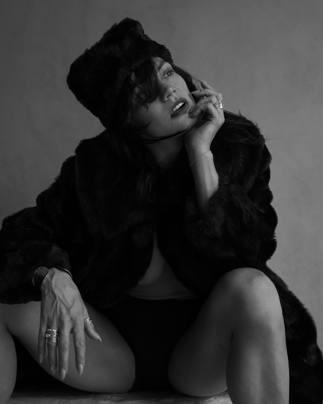 Alisa Pleskova seated in a black fur coat and hat, head tilted, fingers at her lips, black and white