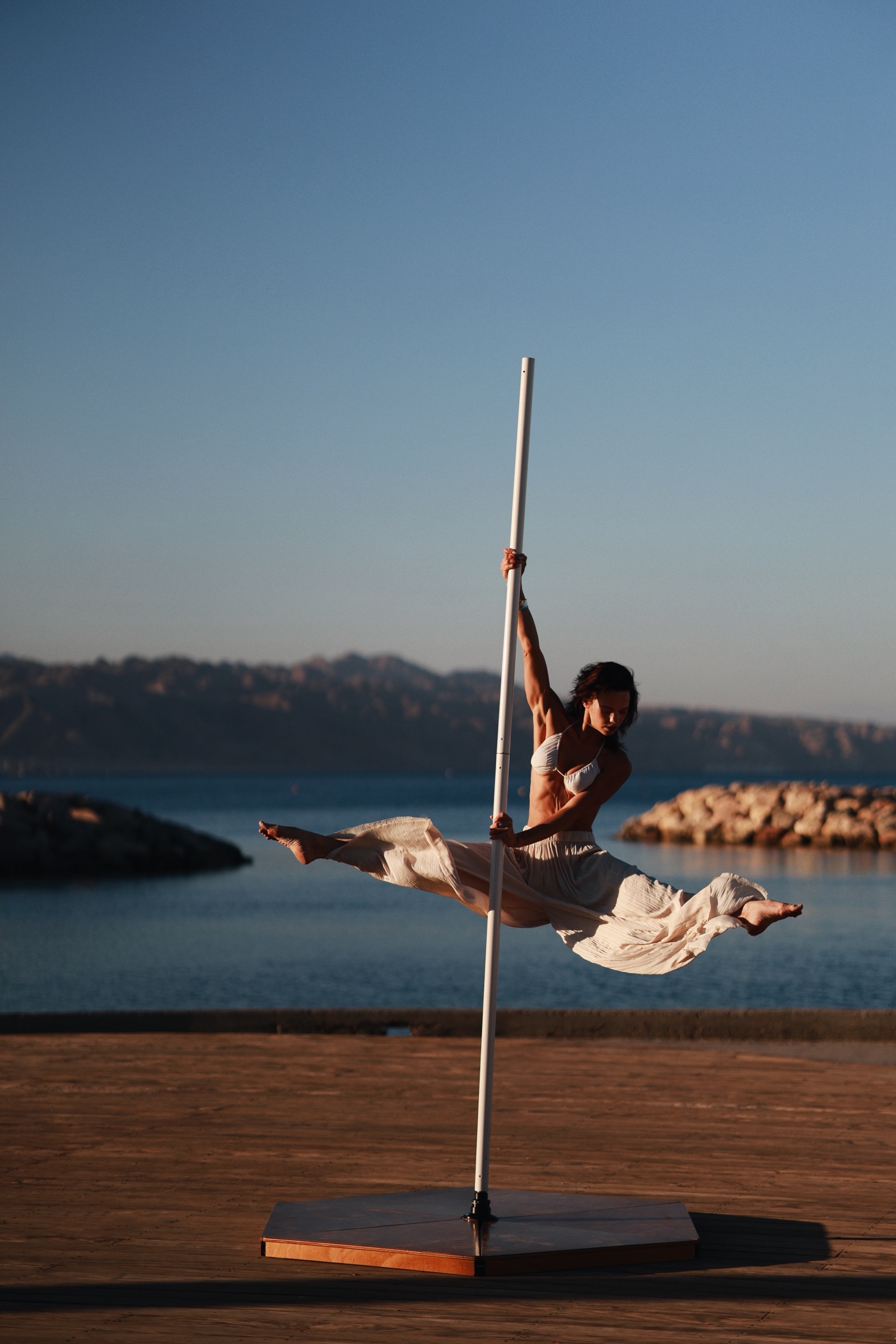 Alisa Pleskova in a split on a pole by the sea at golden hour, white skirt flying